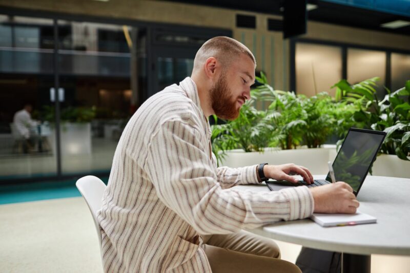 Side,View,Of,Concentrated,Adult,Man,Using,Laptop,And,Writing